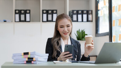 Young attractive Asian business woman with messy desk in office,  sitting at the desk, working at contemporary office and documents. holding coffee cup, using mobile