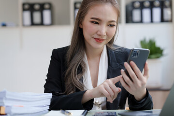 Obraz premium Young attractive Asian business woman with messy desk in office, sitting at the desk, working at contemporary office and documents. texting mobile