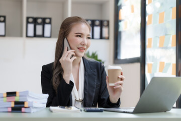 Young attractive Asian business woman with messy desk in office,  sitting at the desk, working at contemporary office and documents. make a mobile phone call