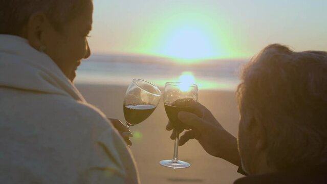 Romantic seniour couple drinking red wine while having picnic on beach at sunset. Back closeup view of clinking glasses and sunset at sea in background. Romance, togetherness, love concept.