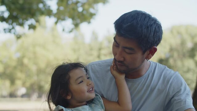 Happy little daughter kissing father on cheek, man taking photo with camera. Young Asian man sitting with toddler girl in park. Fatherhood and childhood concept