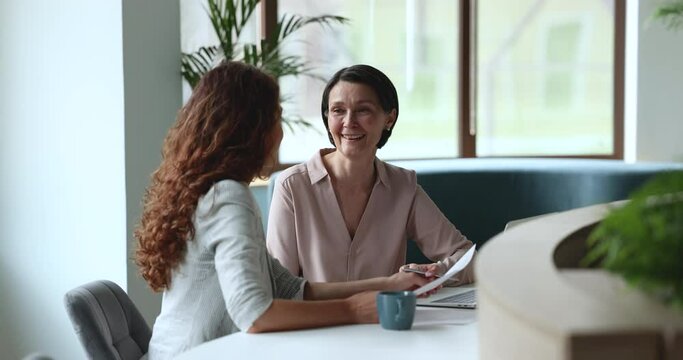 Two attractive businesswomen, middle-aged, millennial client and sales manager sit at desk discuss contract details, discuss agreement or paperwork meet in modern office. Negotiations, deal, commerce