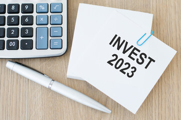 Invest 2023 inscription on a card on a paper clip to a stack of cards on a wooden table next to a pen and calculator