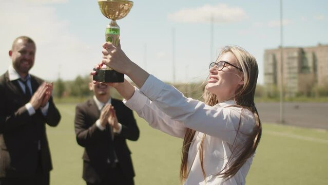 Young caucasian woman celebrates recieving an award for achievement of high work results. Attractive long-hair woman raises winning cup over her head surrounding by cheering colleagues after awarding