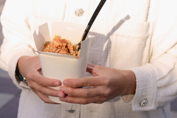 Woman holding paper box of takeaway noodles with fork, closeup. Street food