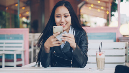 Good-looking Asian girl is making online payment using credit card and smart phone sitting at table in open-air cafe and smiling. Shopping and modern technology concept.