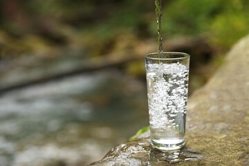 Fresh water pouring into glass on stone near stream. Space for text