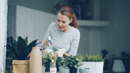 Smiling young woman is spraying green plants with water using spray bottle standing near table in modern loft style apartment. Youth, interior and household concept.