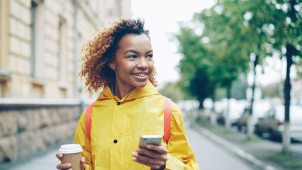 Happy African American woman is using smartphone touching screen and smiling walking outdoors in...