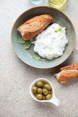 Plate with italian ricotta cheese, green olives and baguette on a beige stone background, view from above, vertical shot