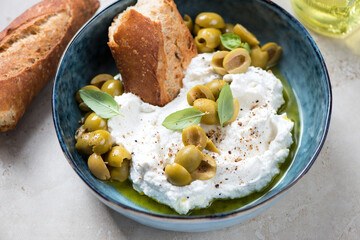 Ricotta cheese served with green olives, olive oil and crusty bread in a blue bowl, middle closeup, horizontal shot