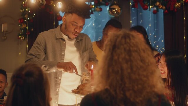 African American man cuts turkey or chicken with knife. Multi cultural friends celebrating Christmas or New Year. Served holiday table with candles. Warm atmosphere of Christmas dinner at home.