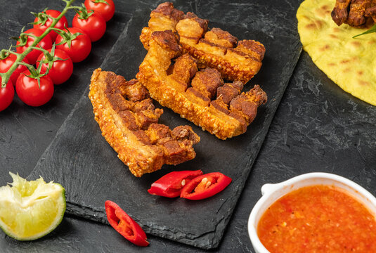 Grilled pork belly with crispy skin. chicharones, a traditional snack. Dark table. Spicy pepper and tomato sauce.