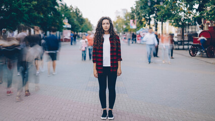 Portrait of beautiful young girl with long curly hair standing in street looking at camera when many men and women are walking around in hurry on summer day. - 549390138