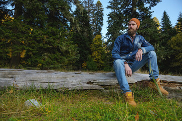 man with a beard sits on a log in the forest. Adventure, travel, tourism and vacation concept - smiling man relaxing in the forest