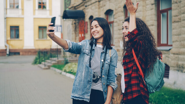 Cheerful girls foreign travelers are taking selfie using smartphone standing outdoors and posing with hand gestures showing v-sign and heart with fingers and laughing together.
