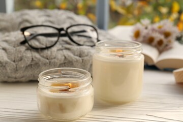 Burning scented candles, warm sweater, glasses and chamomile flowers on white wooden table near window