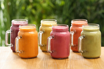 Many different delicious smoothies in mason jars on wooden table against blurred background