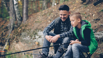 Handsome bearded guy loving dad is teaching his son to catch fish sitting on river bank together, boy is holding rod and moving it talking to daddy. Hobby, adventure and family concept.