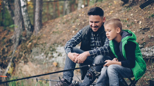 Handsome bearded guy loving dad is teaching his son to catch fish sitting on river bank together, boy is holding rod and moving it talking to daddy. Hobby, adventure and family concept. - Powered by Adobe