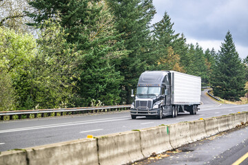 Dark gray big rig professional semi truck transporting cargo in refrigerator semi trailer driving on the straight highway road