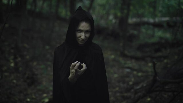 Portrait of a witch in a black cloak with a hood. Woman in the mystical forest. Dark forces