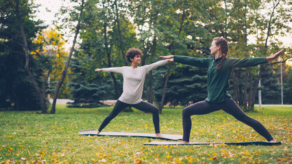 Two beautiful girls yoga teacher and student are practising sequence of asanas in park standing on mats and moving body and arms. Millennials, health and leisure concept.