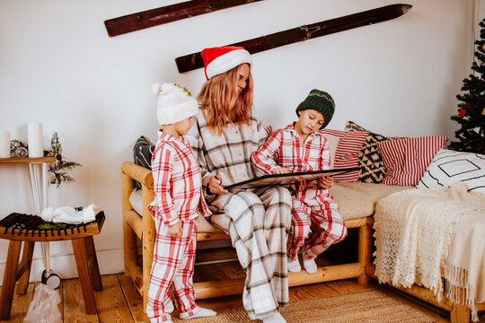 Pretty young woman and little kids in winter pyjamas and hats are drawing and playing together in chalet with Christmas decor. Holiday concept.