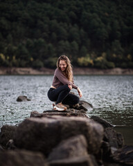 Young beautiful girl sitting on a rock in the middle of a lake with a mountain landscape background smilling at the camera in winter season