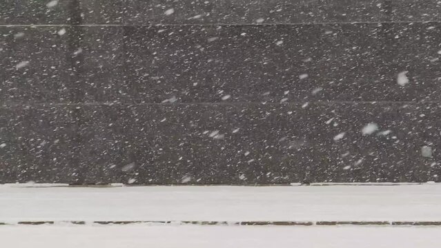 Snow is falling on the street near the wall of the building. A building with a black granite wall, next to a sidewalk and a road covered with white snow, heavy snow falls on the road.