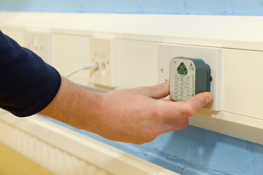Close-up of electrician's hands using electrical circuit tester on UK double wall socket in office