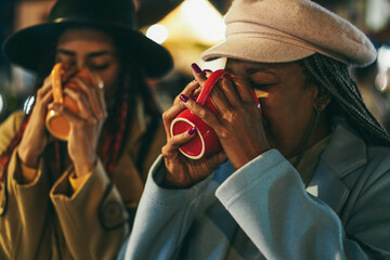 African mother and daughter having fun drinking hot chocolate outdoor in winter time - Focus on senior hands holding cup