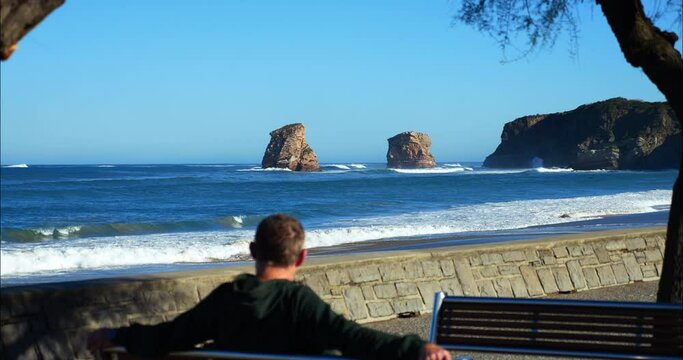 Man sitting on a bench on the Hendaye sea wall with a zoom on the twin rocks