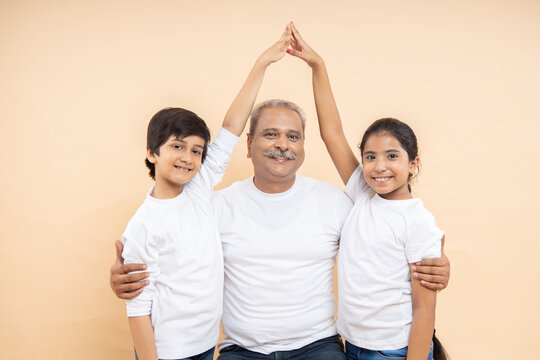 Happy Indian Senior Man With Kids Wearing White Casual T-shirt Make Home Pose With Hand Over Isolated Beige Background.