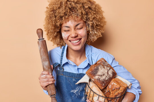 Horizontal shot of positive female baker holds rolling pin and baset full of freshly baked bread works in bakery shop wears blue shirt and denim sarafan enjoys eating organic products poses indoor