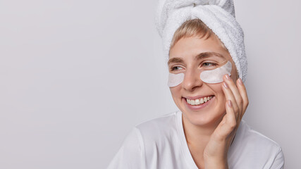 Horizontal shot of pleased European woman with healthy clean skin applies beauty patches under eyes after taking shower looks gladfully shows white teeth isolated over grey background empty space