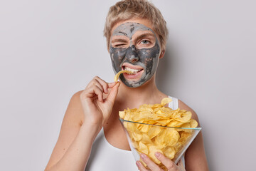 Horizontal shot of European woman with short hair eats delicious crisps enjoys harmful food winks...