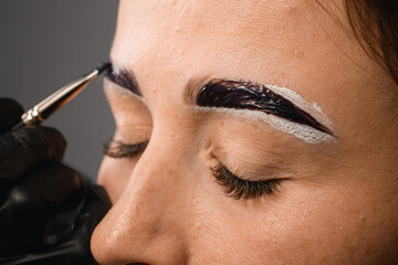 hand in sterile gloves shaping woman eyebrows and applying paint on it using brush. Close-up