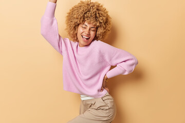People positive emotions and happiness concept. Happy upbeat woman dances carefree feels joy wears knitted jumper trousers keeps arm raised up celebrates at party isolated over beige background