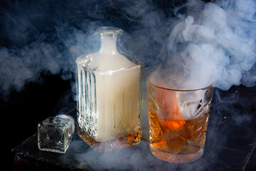 Shrouded in smoke whisky glass decanter with smoke and glass of whiskey with an ice and orange peel...
