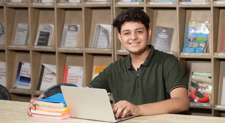 Happy young indian student boy working on laptop and studying in college library with bookshelf behind. working on assignment or project.