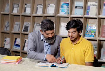 Young indian student boy and mature beard teacher reading book studying in college library with bookshelf behind. - obrazy, fototapety, plakaty