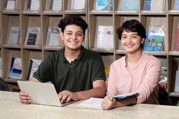 Obraz premium Young indian students reading book and using laptop studying in college library with bookshelf behind. working on assignment or project. together.