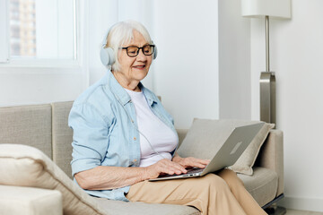 a happy elderly lady is sitting on a cozy sofa in a bright apartment, wearing stylish clothes and holding a laptop on her knees smiling joyfully with her eyes closed listening to music on headphones