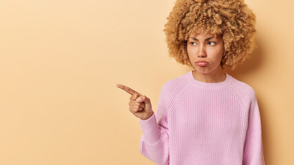 Moody displeased woman expresses regret and disappointment complains about something bad indicates fore finger aside wears pink knitted sweater isolated over beige background tells upsetting news