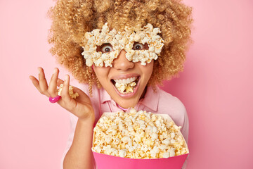 Image of thrilled glad woman has mouth full of popcorn eats delicious snack while watching movie in...