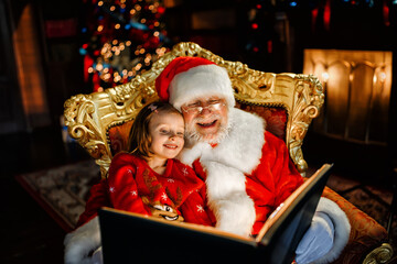 Fototapeta na wymiar Santa Claus is sitting in a chair with a child on his lap and reading a book