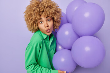 Surprised curly woman prepares for party decorates room holds bunch of inflated balloons dressed in green jumper reacts to something shocking isolated over purple background. Celebration concept