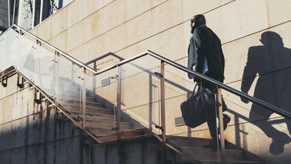 A bald black man with a beard walks up the stairs to work. The successful African male is ascending a staircase on a sunny day, carrying a notebook and a leather bag wearing a grey suit and sunglasses