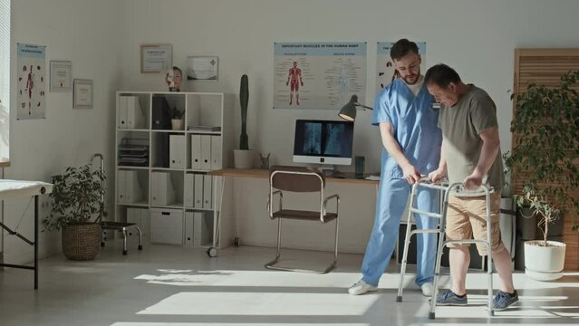 Young therapist helping mature patient to walk with walker during his rehabilitation at hospital